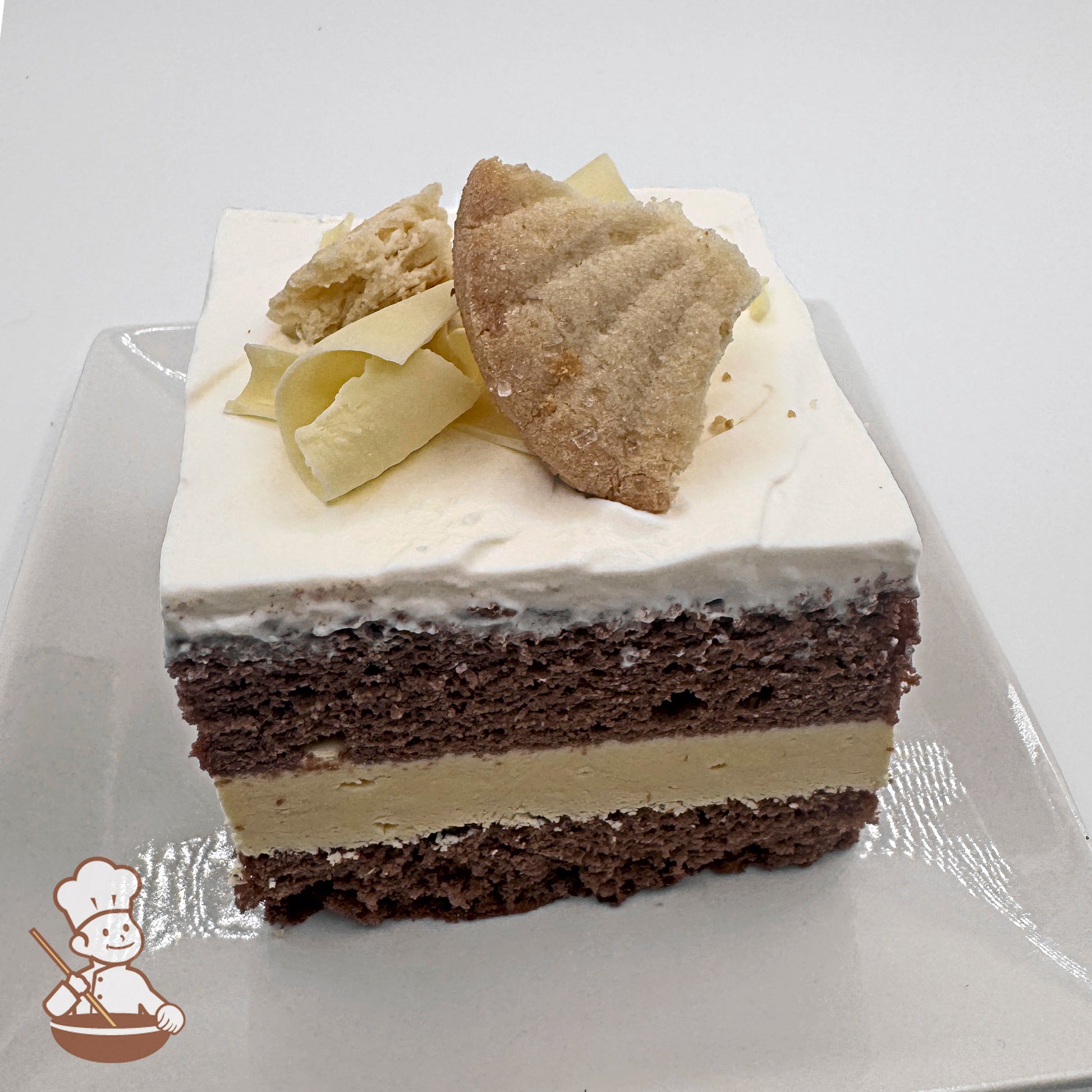 A slice of chocolate chiffon cake topped with white frosting, white chocolate curls, and a shortbread cookie on a white plate.