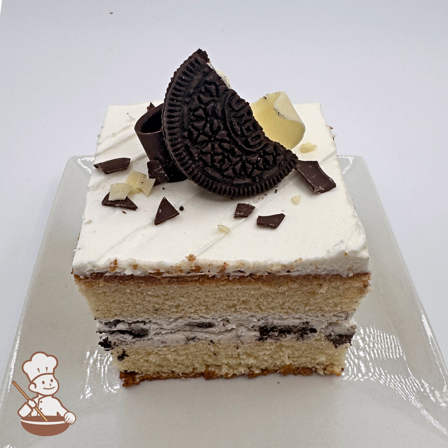 A slice of chocolate chiffon cake topped with buttercream, Oreo pieces, and white chocolate curls, presented on a white plate.