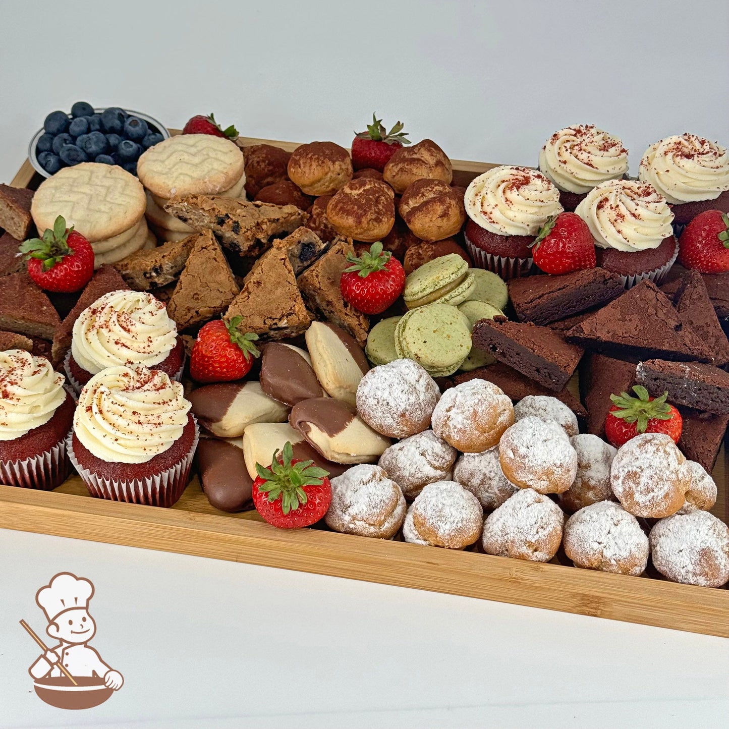 A rectangular bamboo tray carrying red velvet cupcakes, cream puffs, blondies, brownies, shortbreads, a bowl of blueberries, and strawberries.