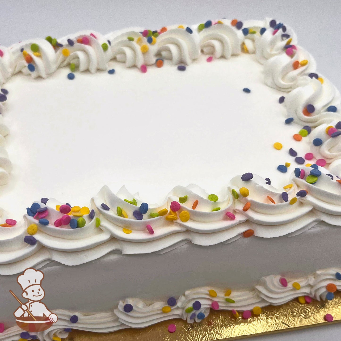 A rectangle cake with white frosting, decorated with colorful sprinkles and piped swirls along the edges, sits on a gold board.