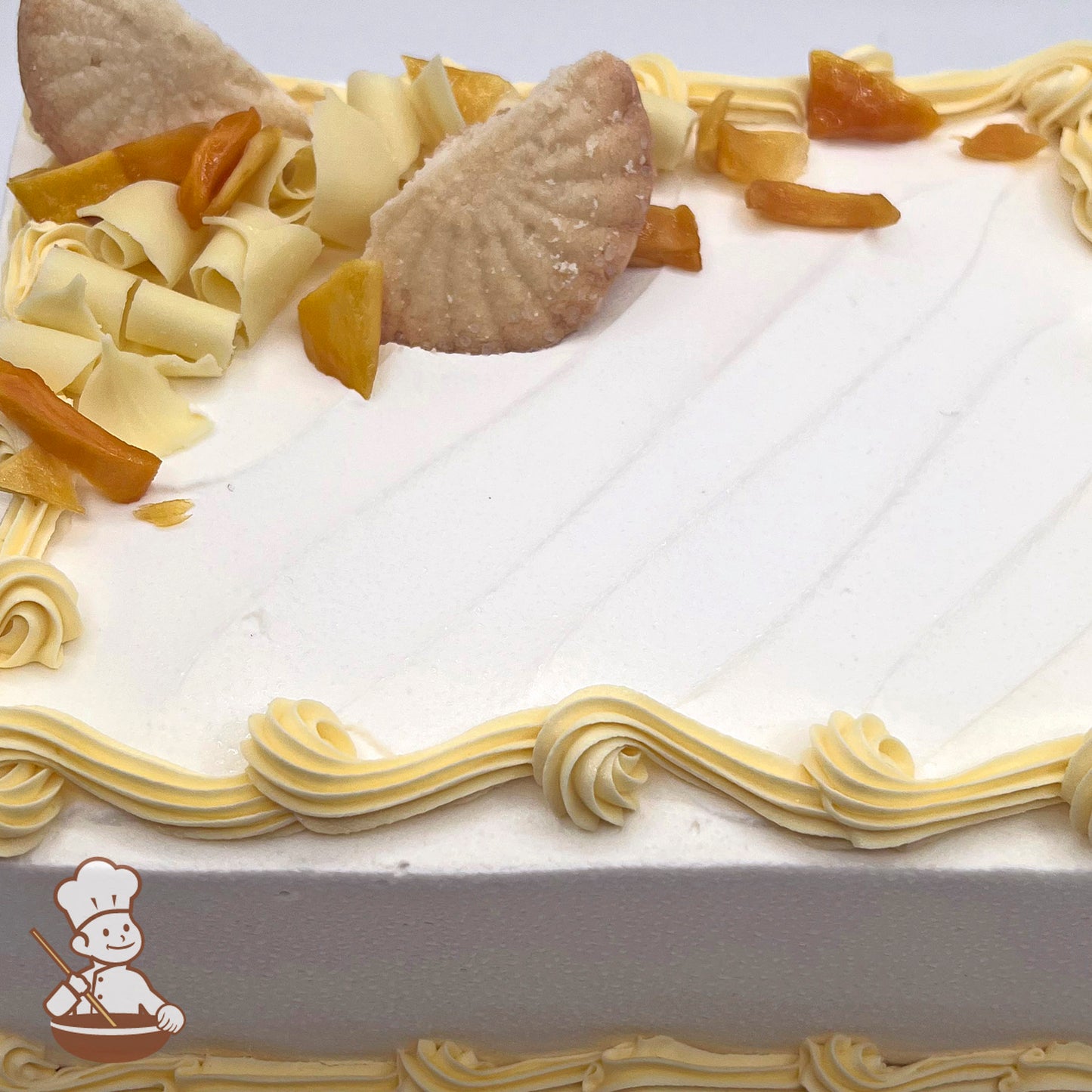 A sheet cake with white frosting, decorated with white chocolate curls, butter shortbread cookies, and dried mango pieces. Creamy swirls edge the cake.