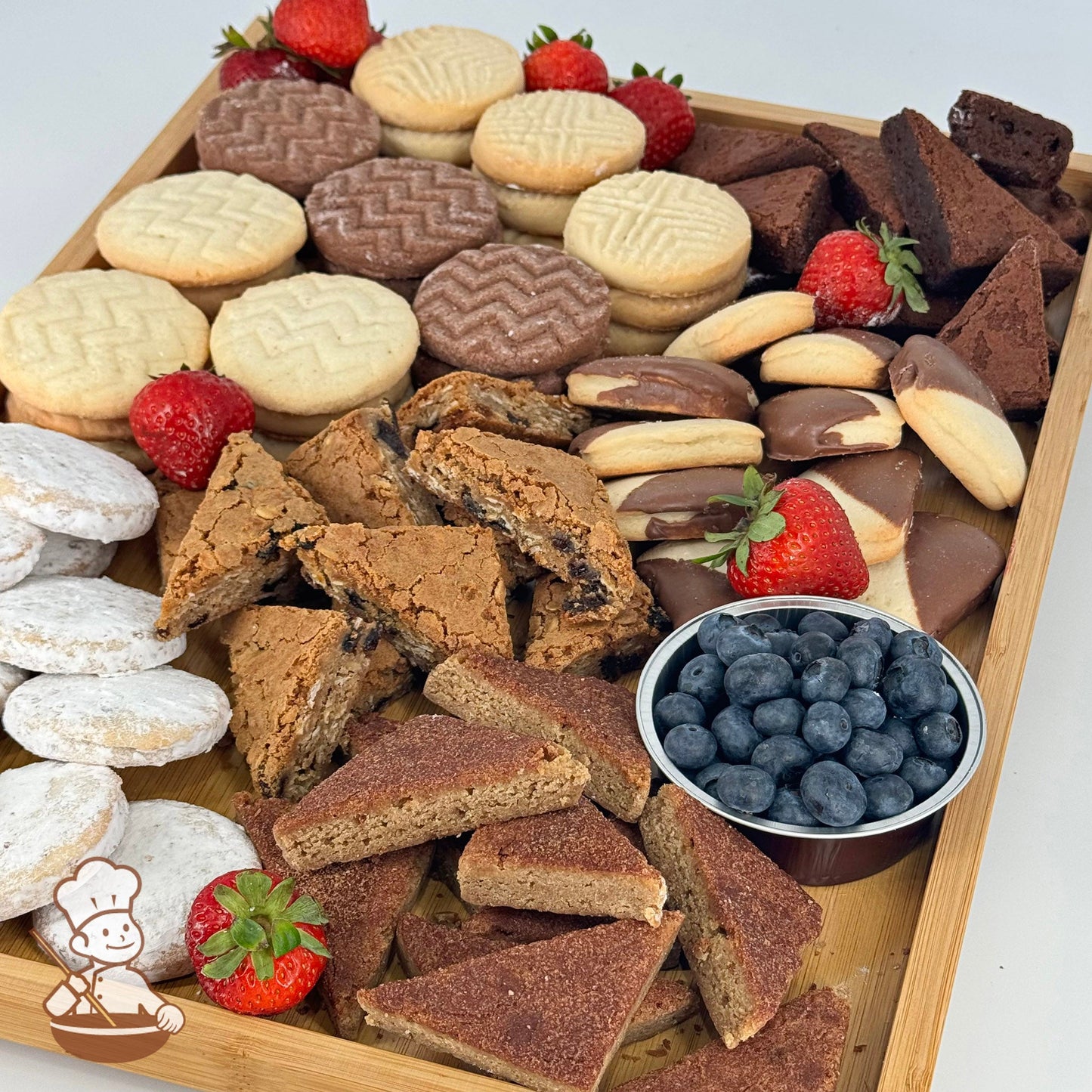 A rectangular bamboo tray carrying an assortment of cookies and brownies, fresh blueberries in a bowl, and strawberries.