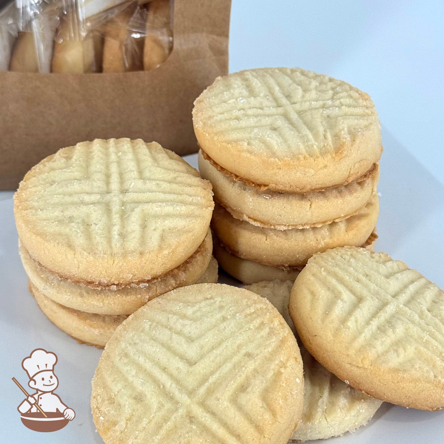 Ten buttery shortbread cookies with a light, airy texture and topped with sugar crystals are stacked in front of a kraft window bag.