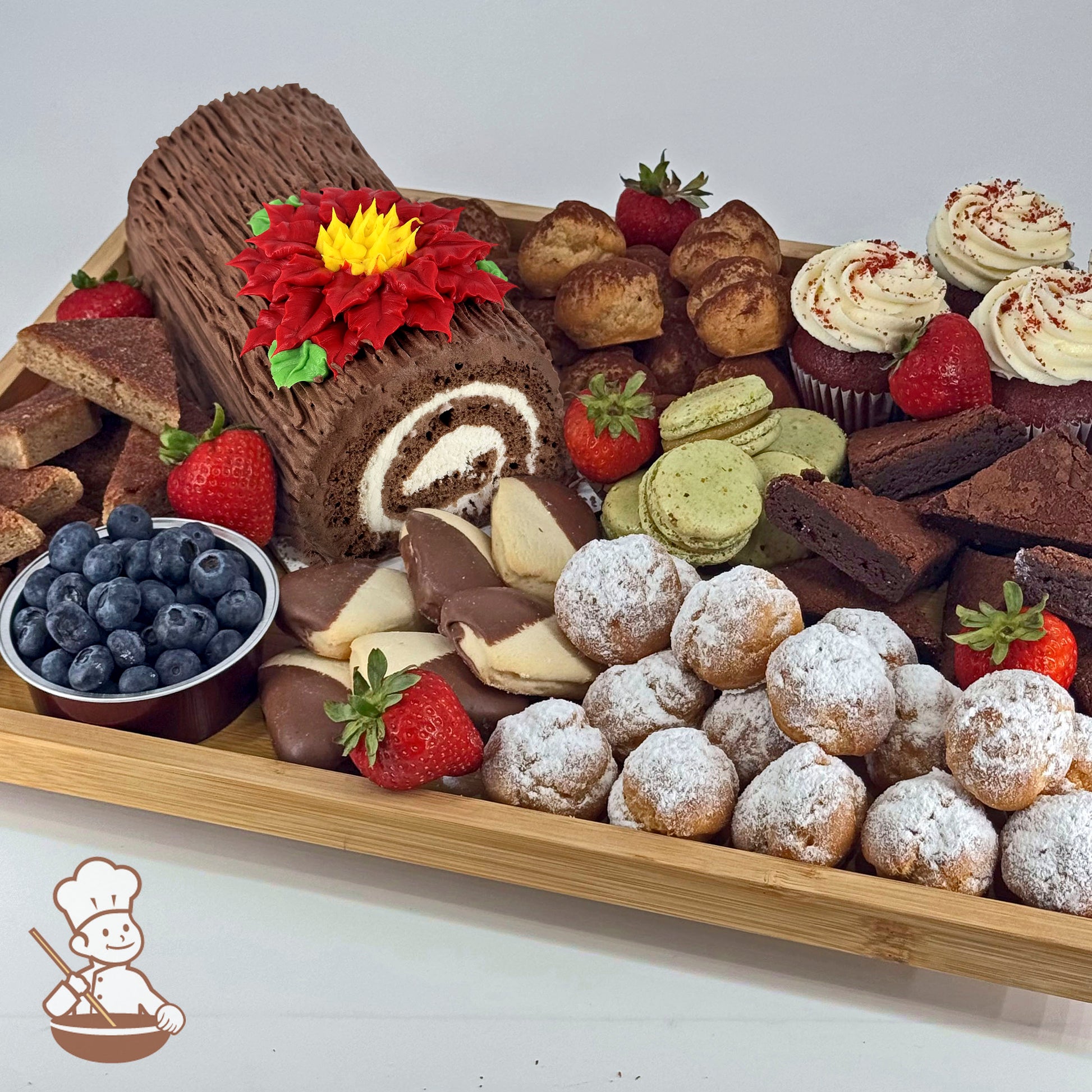 Yule log dessert board with cream puffs, brownies, chocolate-dipped shortbread, red velvet cupcakes, pistachio macarons, strawberries, and blueberries.