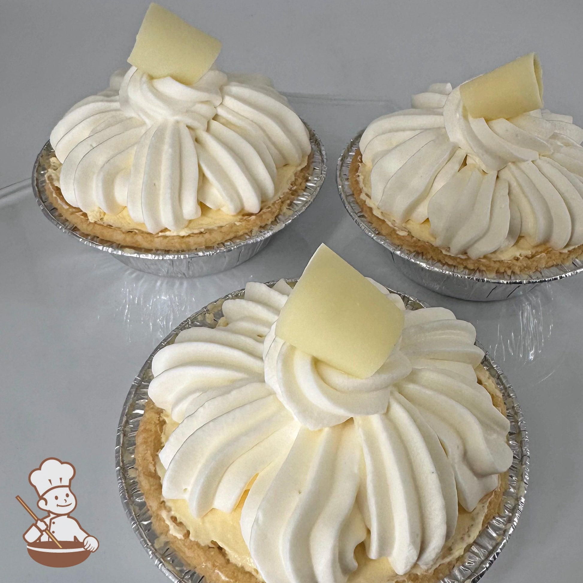 Three mini pies in foil tins with banana cream filling, topped with whip cream swirls and garnished with a white chocolate curl.