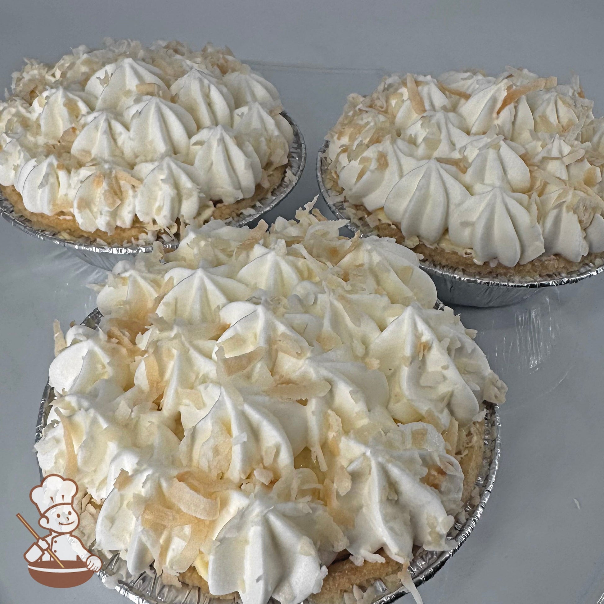 Three mini pies in foil tins filled with Bavarian cream and topped with whipped cream and toasted coconut shavings.