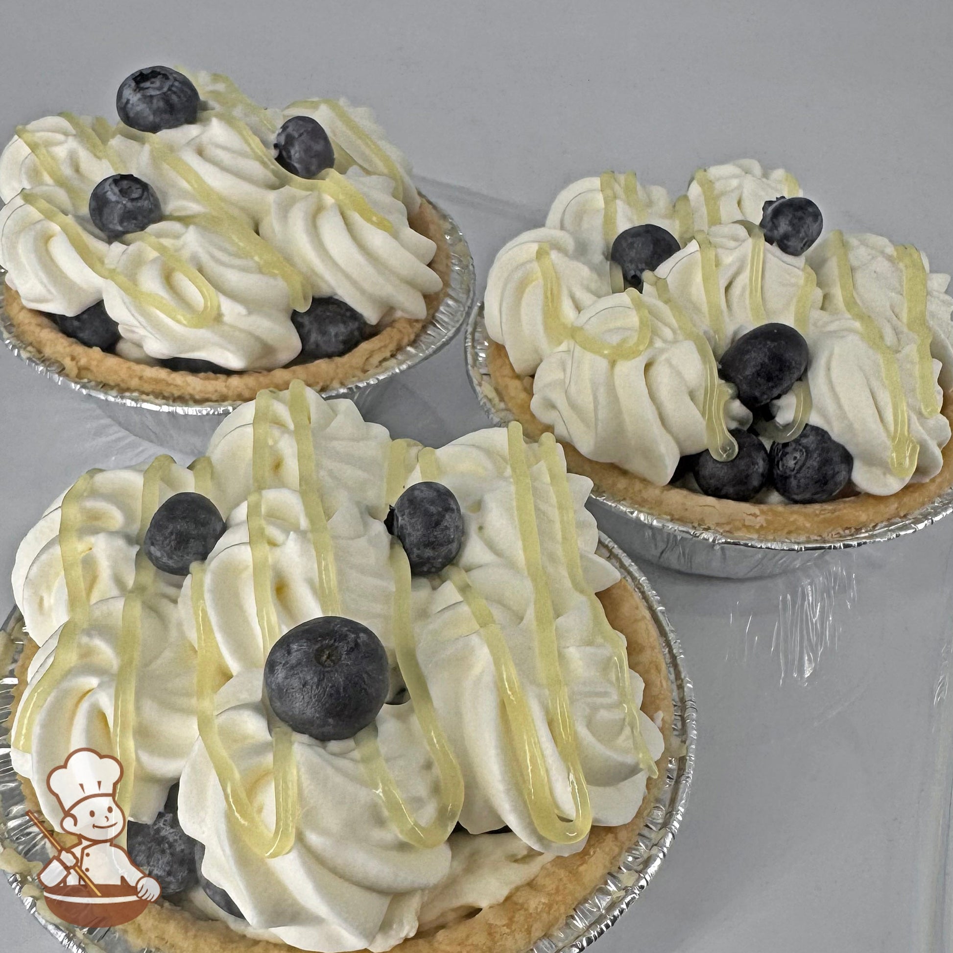 Three mini pies in foil tins with lemon cream filling and whip cream topping, covered with lemon curd drizzle and fresh blueberries.