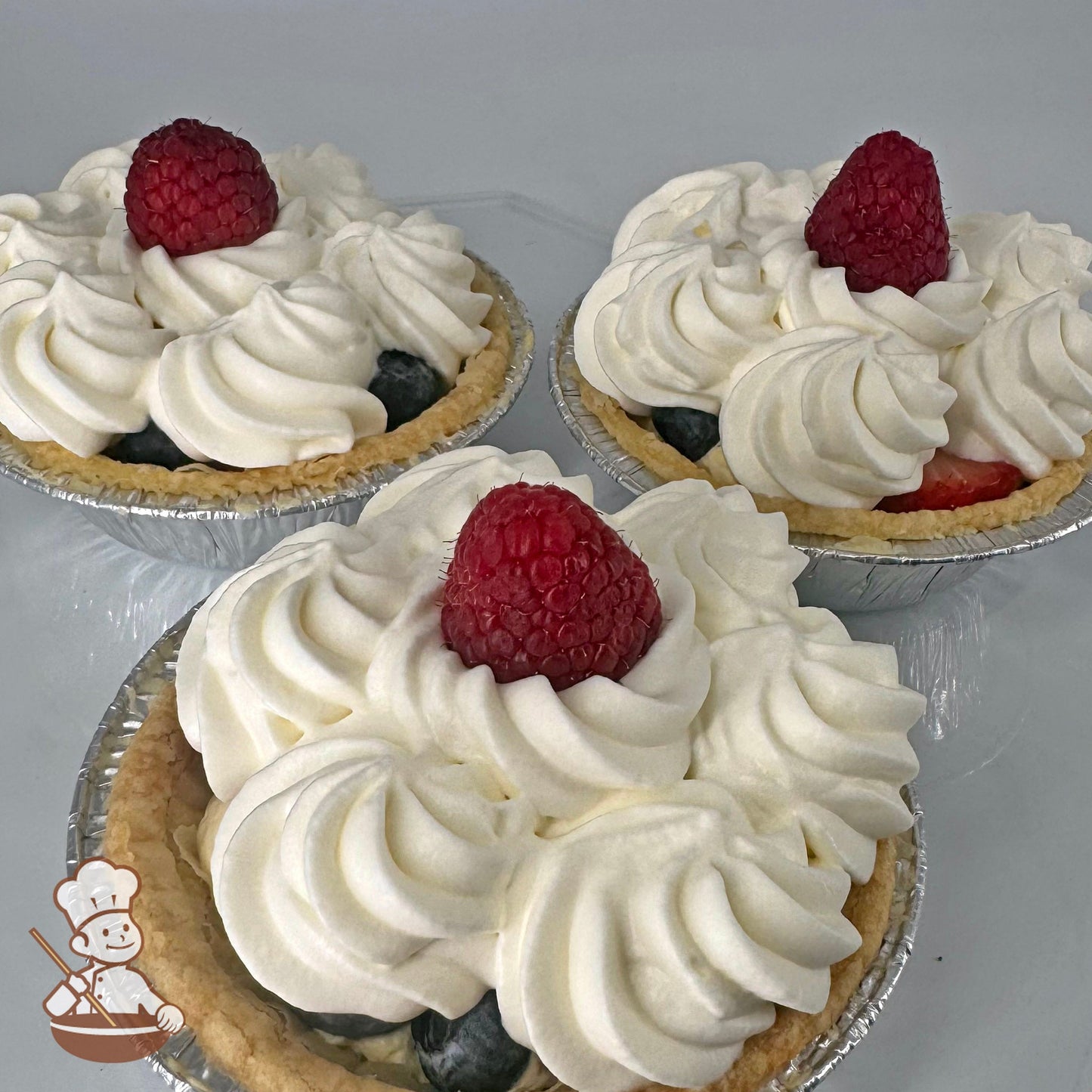 Three mini pies in foil tins with Bavarian cream filling, fresh strawberry slices, fresh blueberries, whip cream topping, and a fresh raspberry garnish.