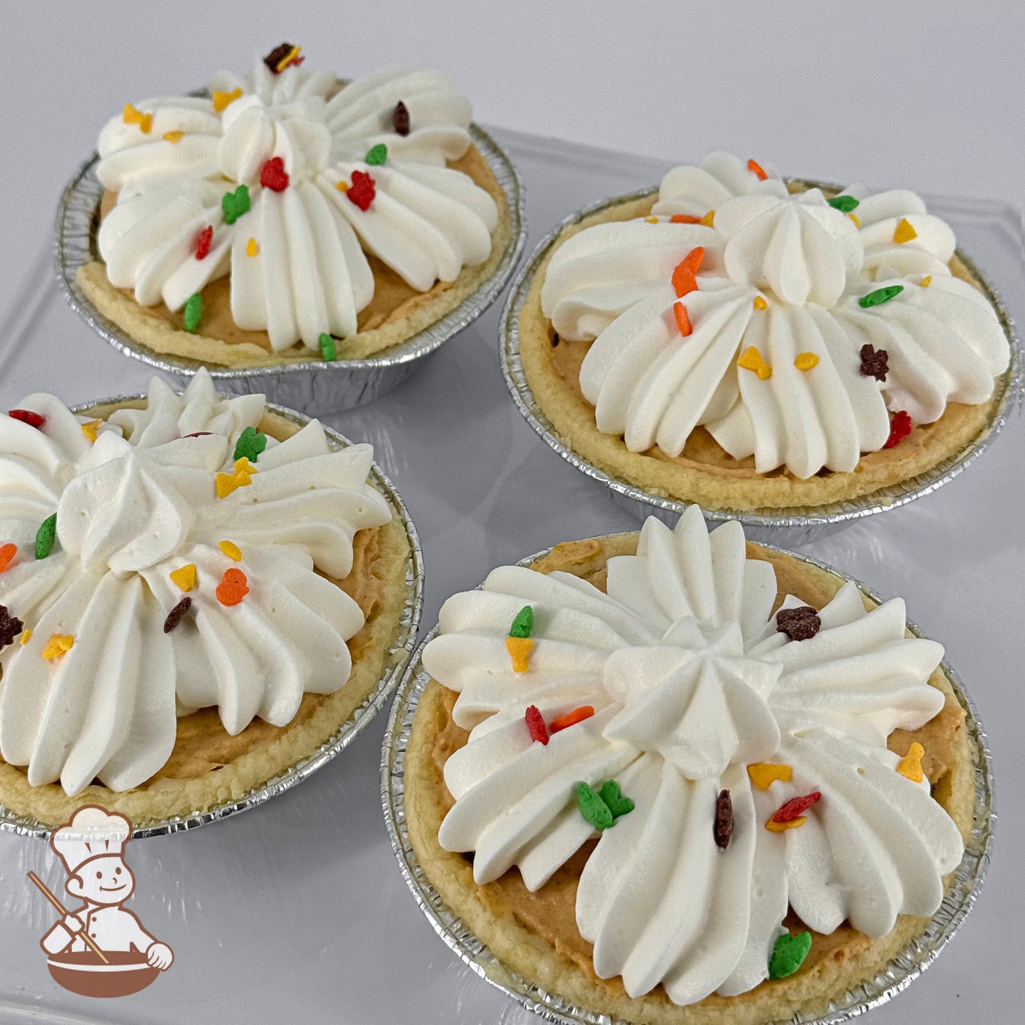 Three mini pumpkin cream pies in foil tins with swirled whipped cream topping and vibrant autumn leaf sprinkles.