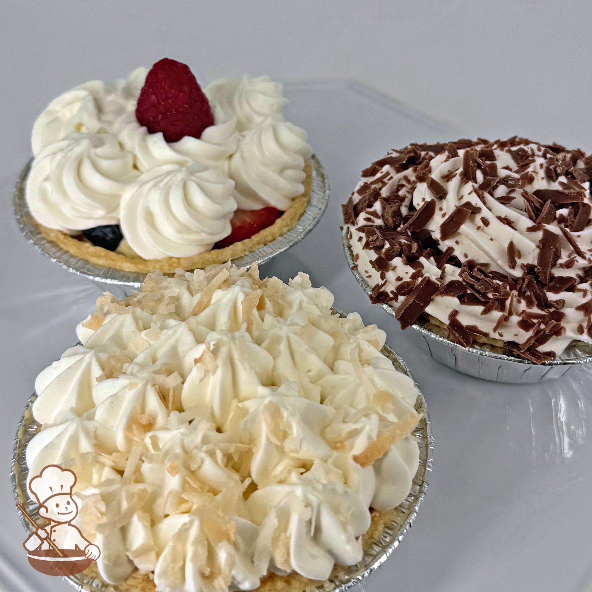 Three mini pies in foil tins, each with different toppings: one with whipped cream and a raspberry, one with chocolate shavings, and one with whipped cream and coconut flakes.