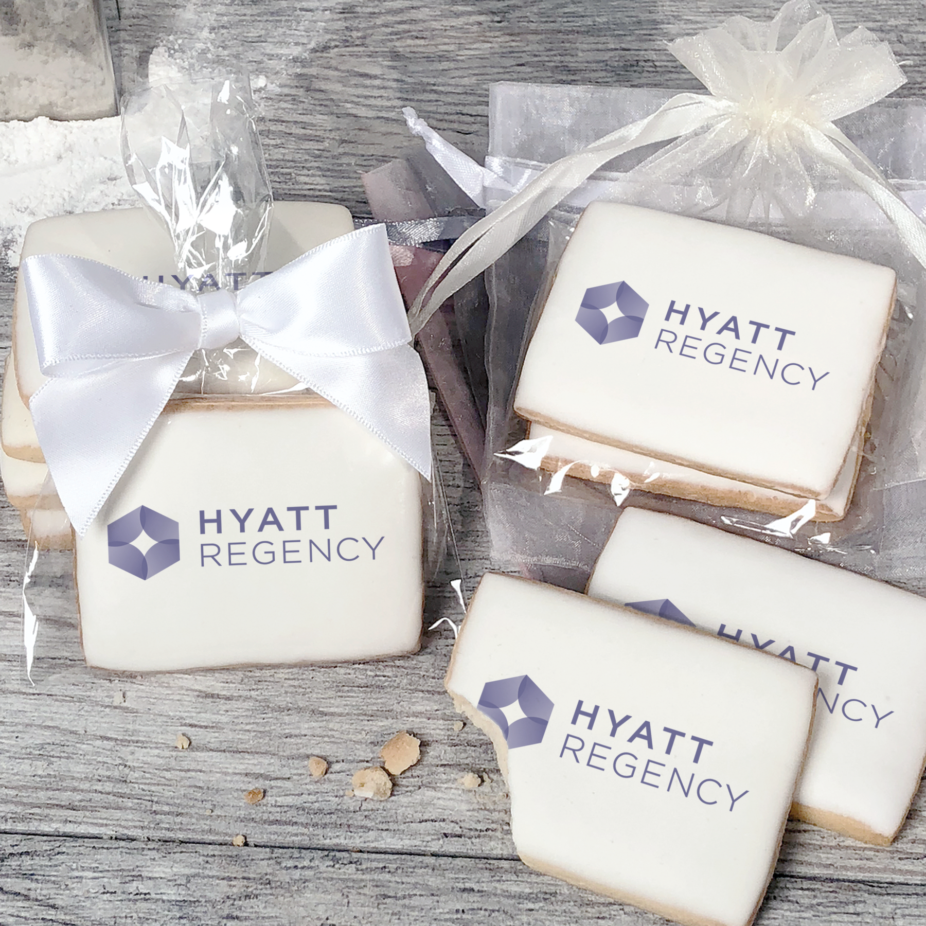 A stack of rectangular butter shortbread cookies with Hyatt Regency logo printed directly on a white, light sugar icing. Some cookies are shown in clear packaging with a twist-tie ribbon bow or inside an organza bag.