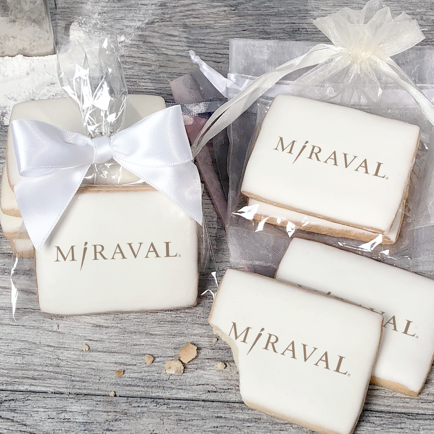 A stack of rectangular butter shortbread cookies with MiRaval logo printed directly on a white, light sugar icing. Some cookies are shown in clear packaging with a twist-tie ribbon bow or inside an organza bag.