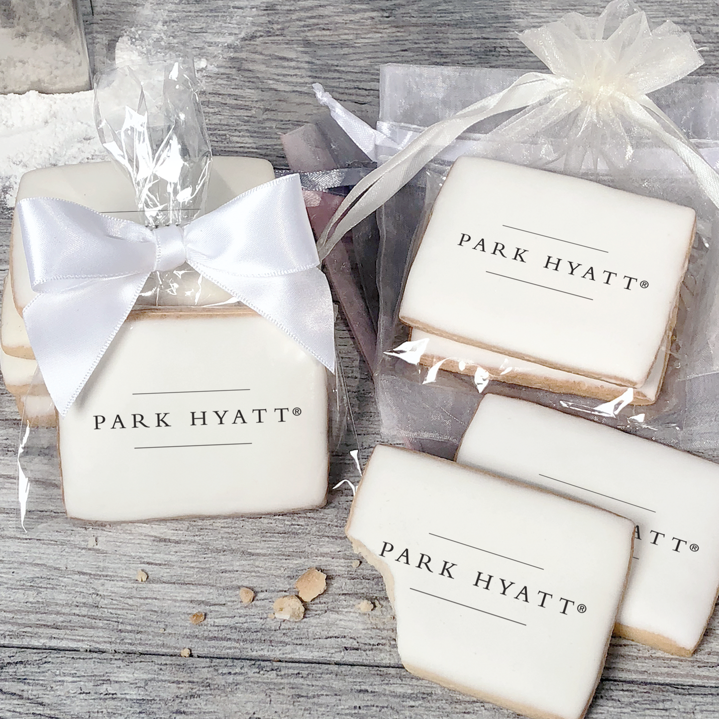 A stack of rectangular butter shortbread cookies with Park Hyatt logo printed directly on a white, light sugar icing. Some cookies are shown in clear packaging with a twist-tie ribbon bow or inside an organza bag.