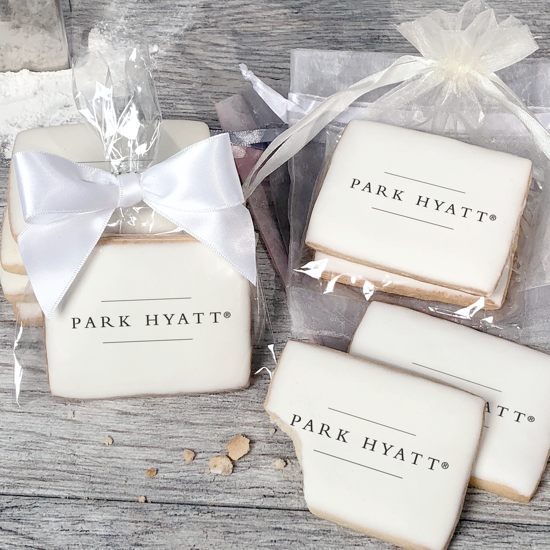 A stack of rectangular butter shortbread cookies with Park Hyatt logo printed directly on a white, light sugar icing. Some cookies are shown in clear packaging with a twist-tie ribbon bow or inside an organza bag.