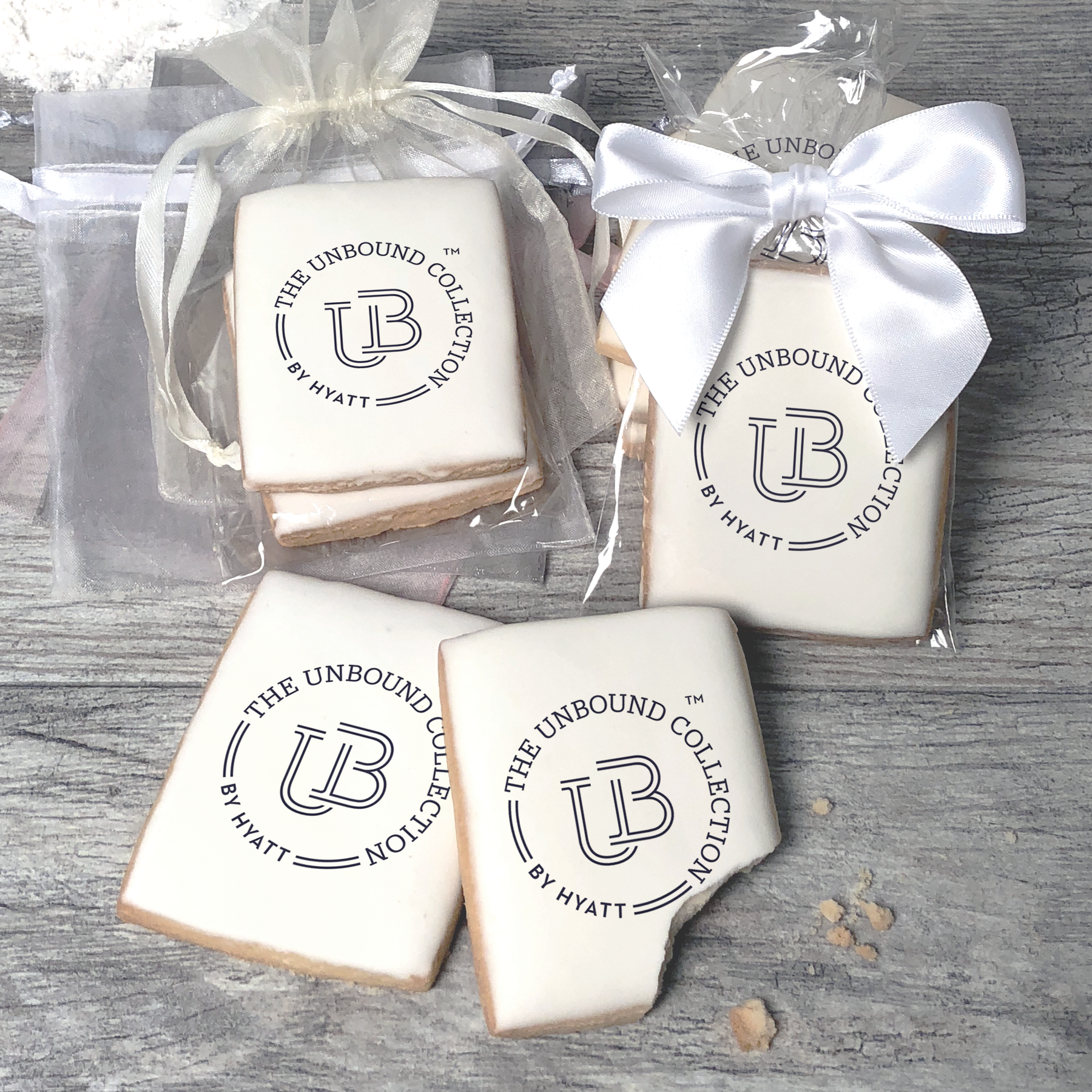 A stack of rectangular butter shortbread cookies with The Unbound Collection by Hyatt logo printed directly on a white, light sugar icing. Some cookies are shown in clear packaging with a twist-tie ribbon bow or inside an organza bag.