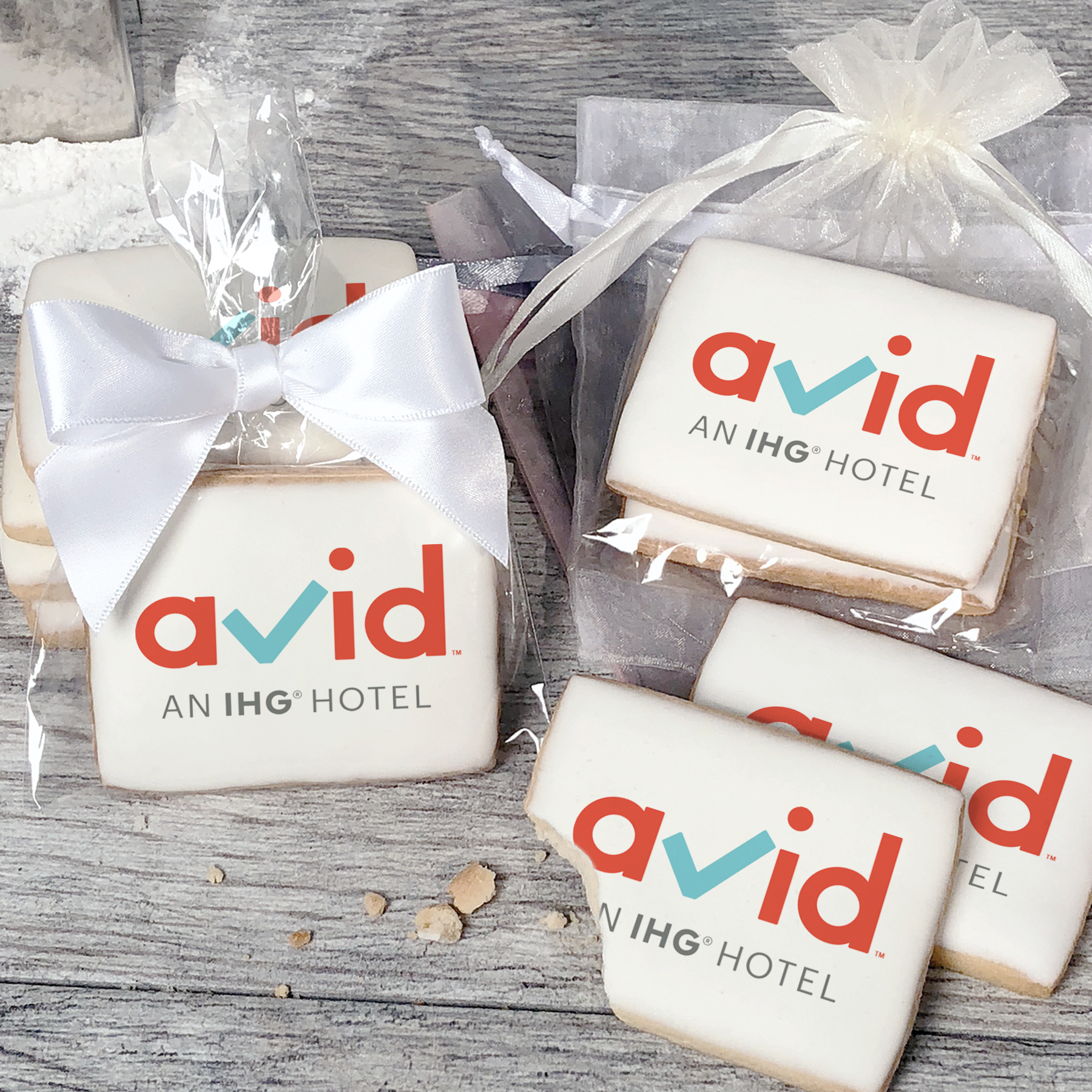 A stack of rectangular butter shortbread cookies with Avid logo printed directly on a white, light sugar icing. Some cookies are shown in clear packaging with a twist-tie ribbon bow or inside an organza bag.