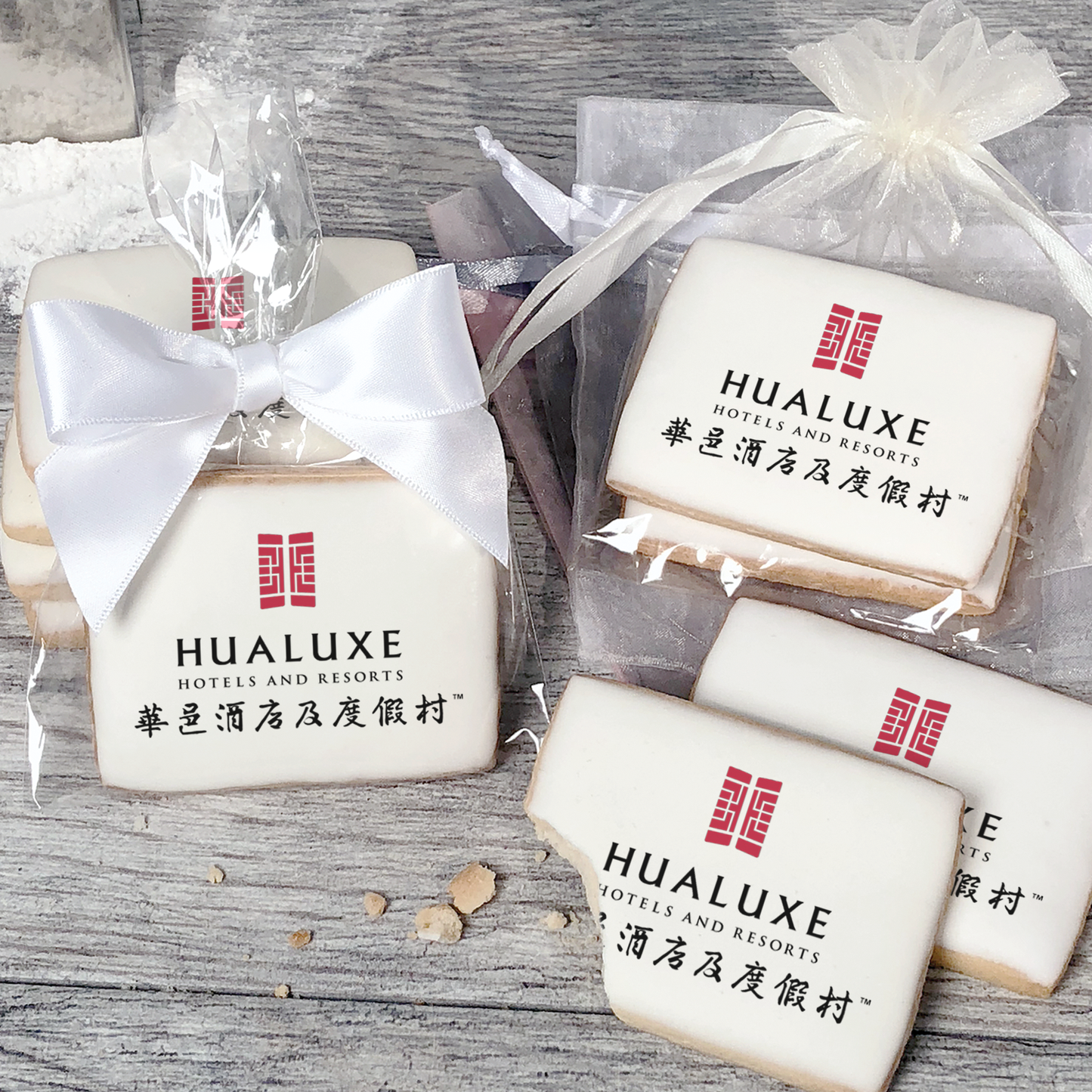 A stack of rectangular butter shortbread cookies with Hualuxe logo printed directly on a white, light sugar icing. Some cookies are shown in clear packaging with a twist-tie ribbon bow or inside an organza bag.