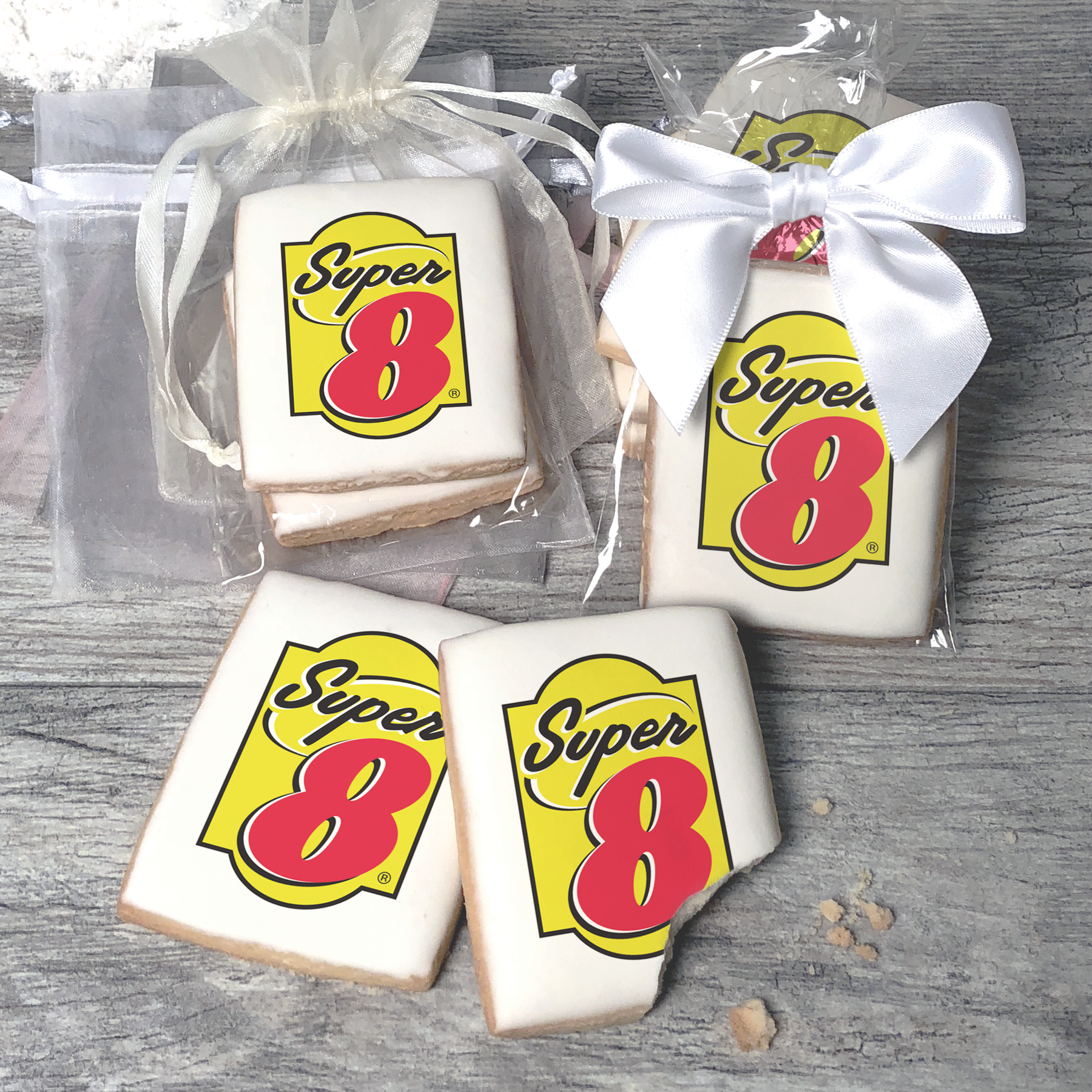 A stack of rectangular butter shortbread cookies with Super 8 logo printed directly on a white, light sugar icing. Some cookies are shown in clear packaging with a twist-tie ribbon bow or inside an organza bag.