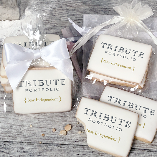 A stack of rectangular butter shortbread cookies with Tribute Portfolio logo printed directly on a white, light sugar icing. Some cookies are shown in clear packaging with a twist-tie ribbon bow or inside an organza bag.