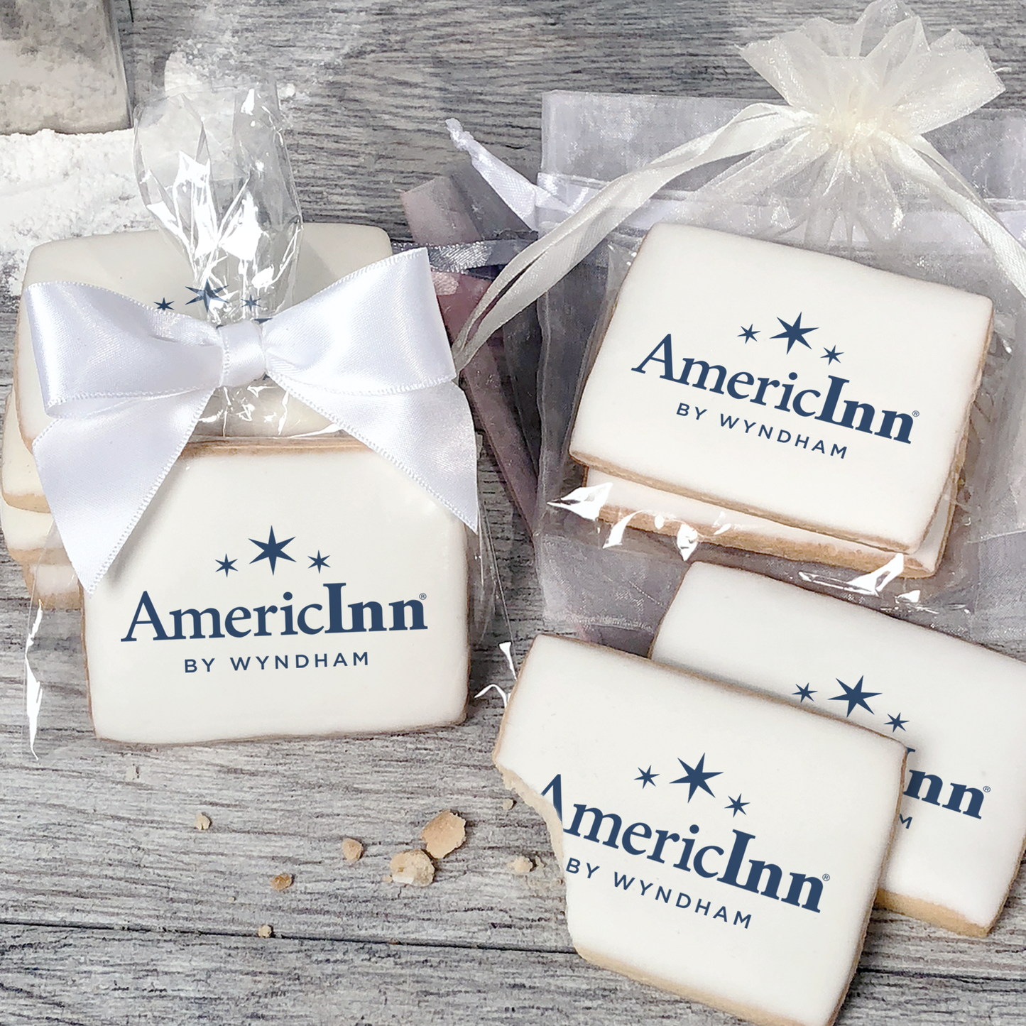 A stack of rectangular butter shortbread cookies with AmericInn logo printed directly on a white, light sugar icing. Some cookies are shown in clear packaging with a twist-tie ribbon bow or inside an organza bag.