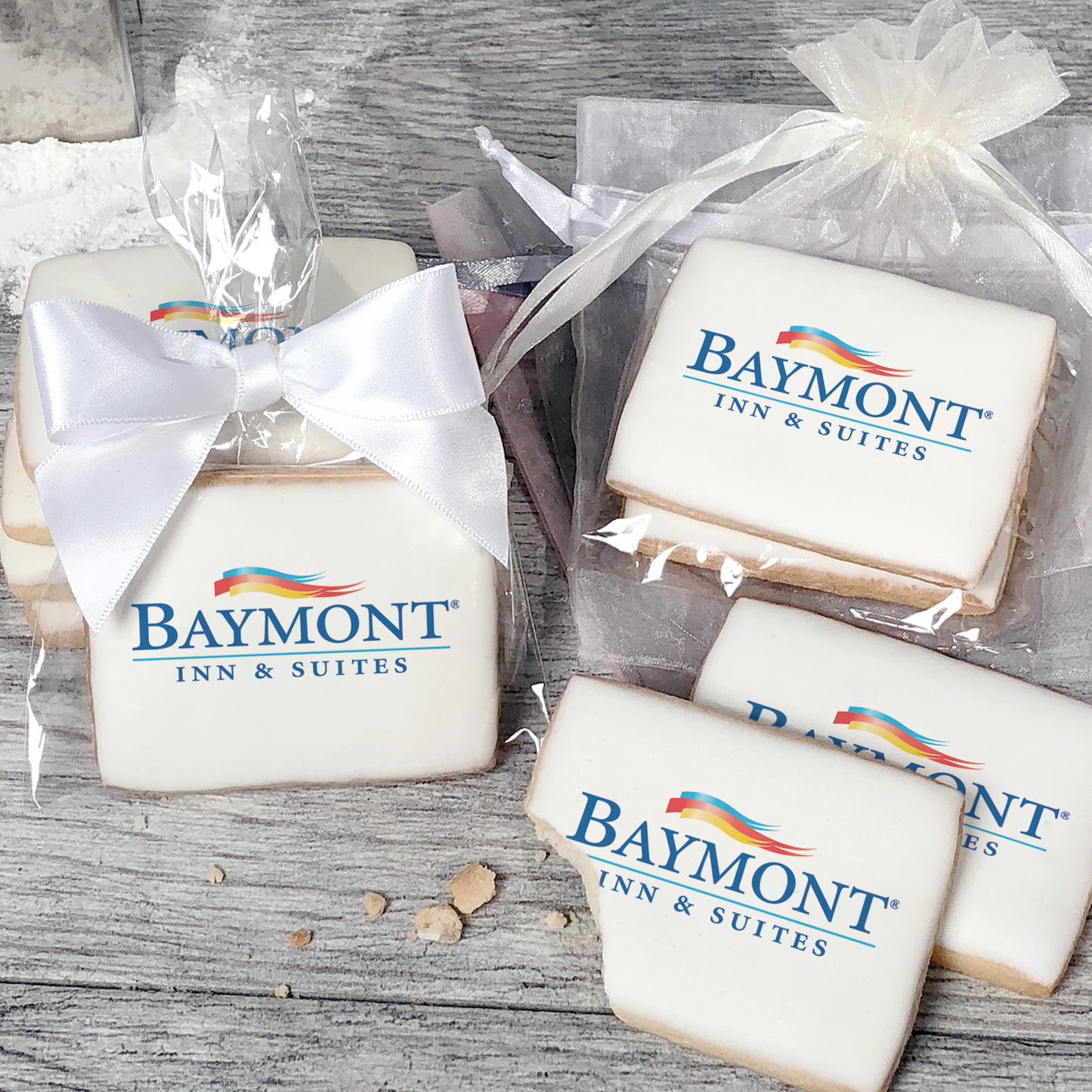 A stack of rectangular butter shortbread cookies with Baymont logo printed directly on a white, light sugar icing. Some cookies are shown in clear packaging with a twist-tie ribbon bow or inside an organza bag.