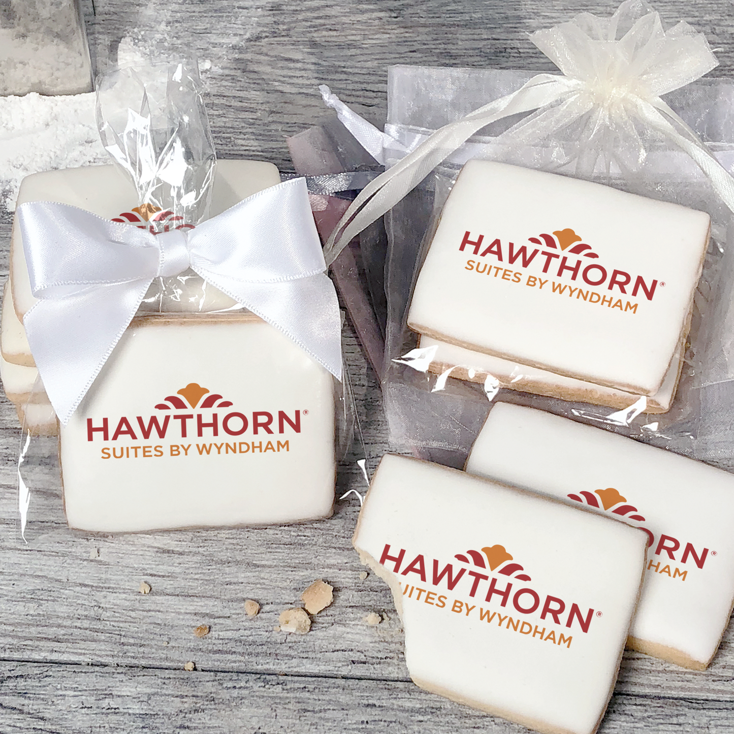 A stack of rectangular butter shortbread cookies with Hawthorn logo printed directly on a white, light sugar icing. Some cookies are shown in clear packaging with a twist-tie ribbon bow or inside an organza bag.