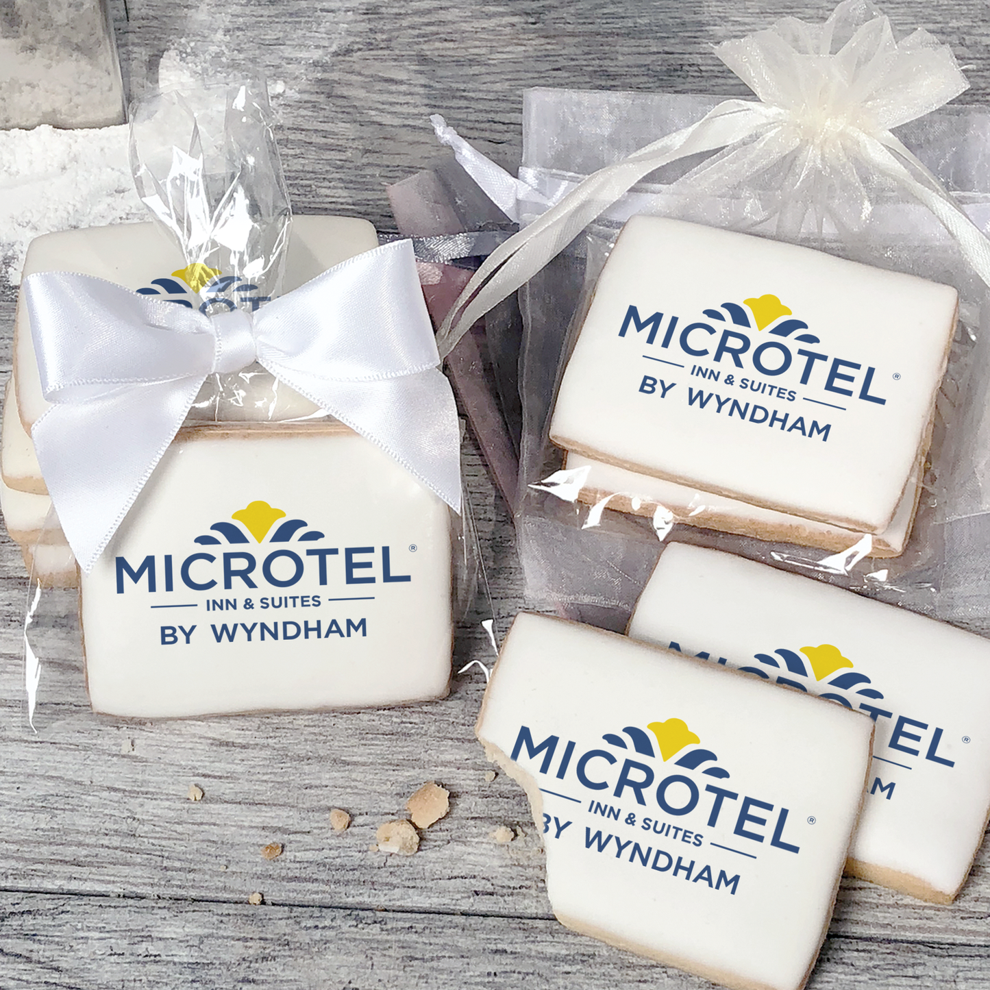 A stack of rectangular butter shortbread cookies with Microtel logo printed directly on a white, light sugar icing. Some cookies are shown in clear packaging with a twist-tie ribbon bow or inside an organza bag.