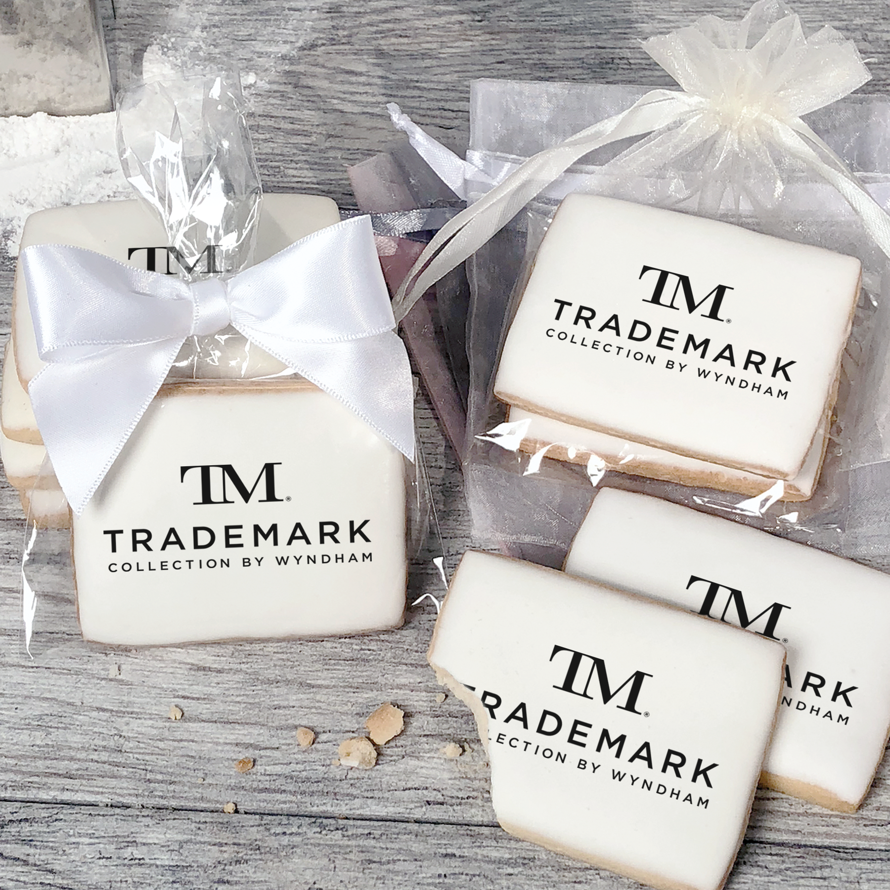 A stack of rectangular butter shortbread cookies with TM Trademark Collection by Wyndham logo printed directly on a white, light sugar icing. Some cookies are shown in clear packaging with a twist-tie ribbon bow or inside an organza bag.