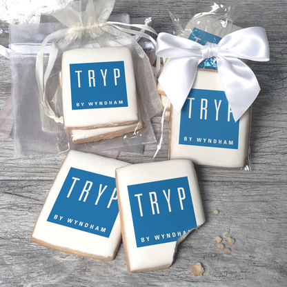 A stack of rectangular butter shortbread cookies with Tryp logo printed directly on a white, light sugar icing. Some cookies are shown in clear packaging with a twist-tie ribbon bow or inside an organza bag.