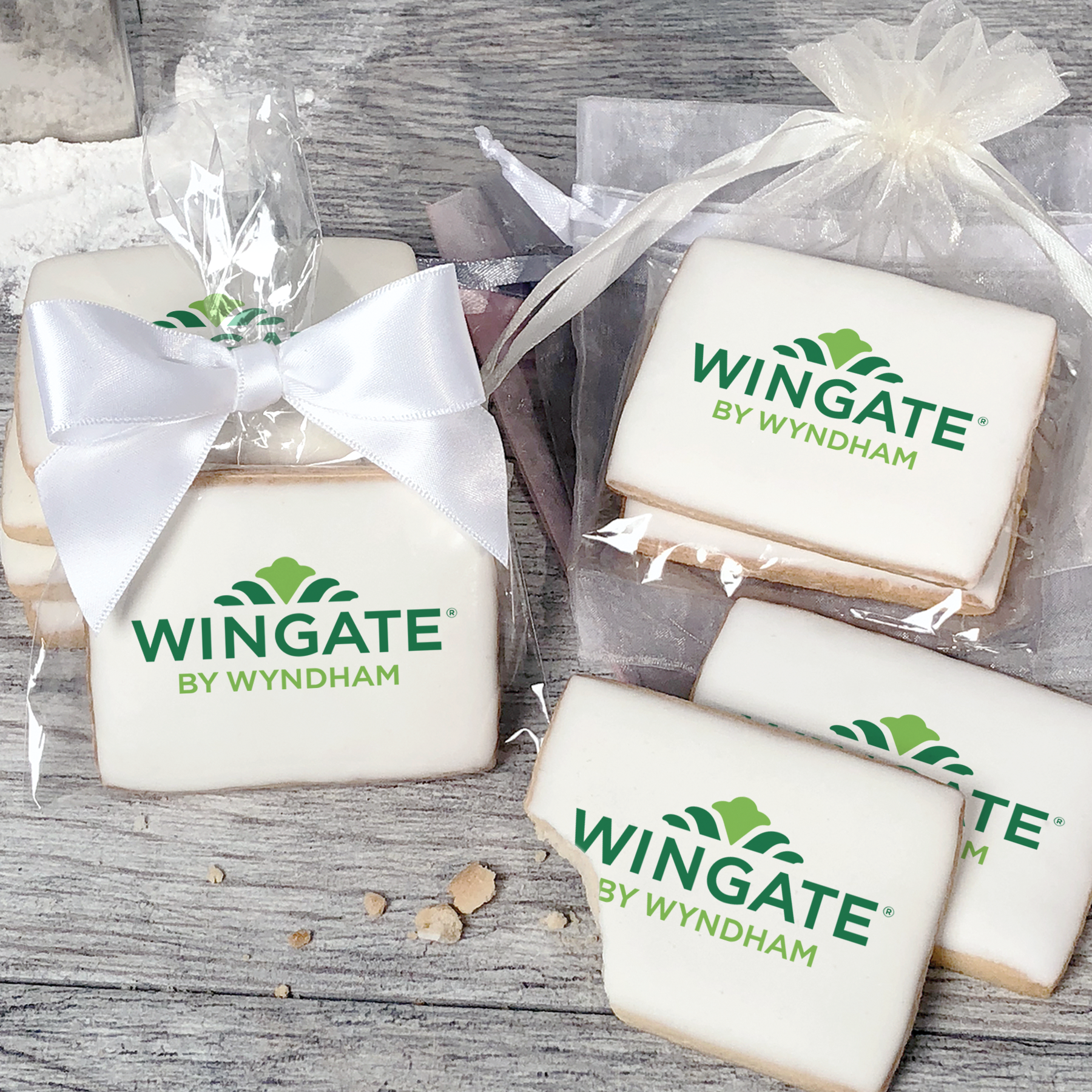 A stack of rectangular butter shortbread cookies with Wingate by Wyndham logo printed directly on a white, light sugar icing. Some cookies are shown in clear packaging with a twist-tie ribbon bow or inside an organza bag.