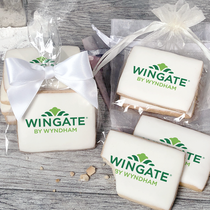 A stack of rectangular butter shortbread cookies with Wingate by Wyndham logo printed directly on a white, light sugar icing. Some cookies are shown in clear packaging with a twist-tie ribbon bow or inside an organza bag.