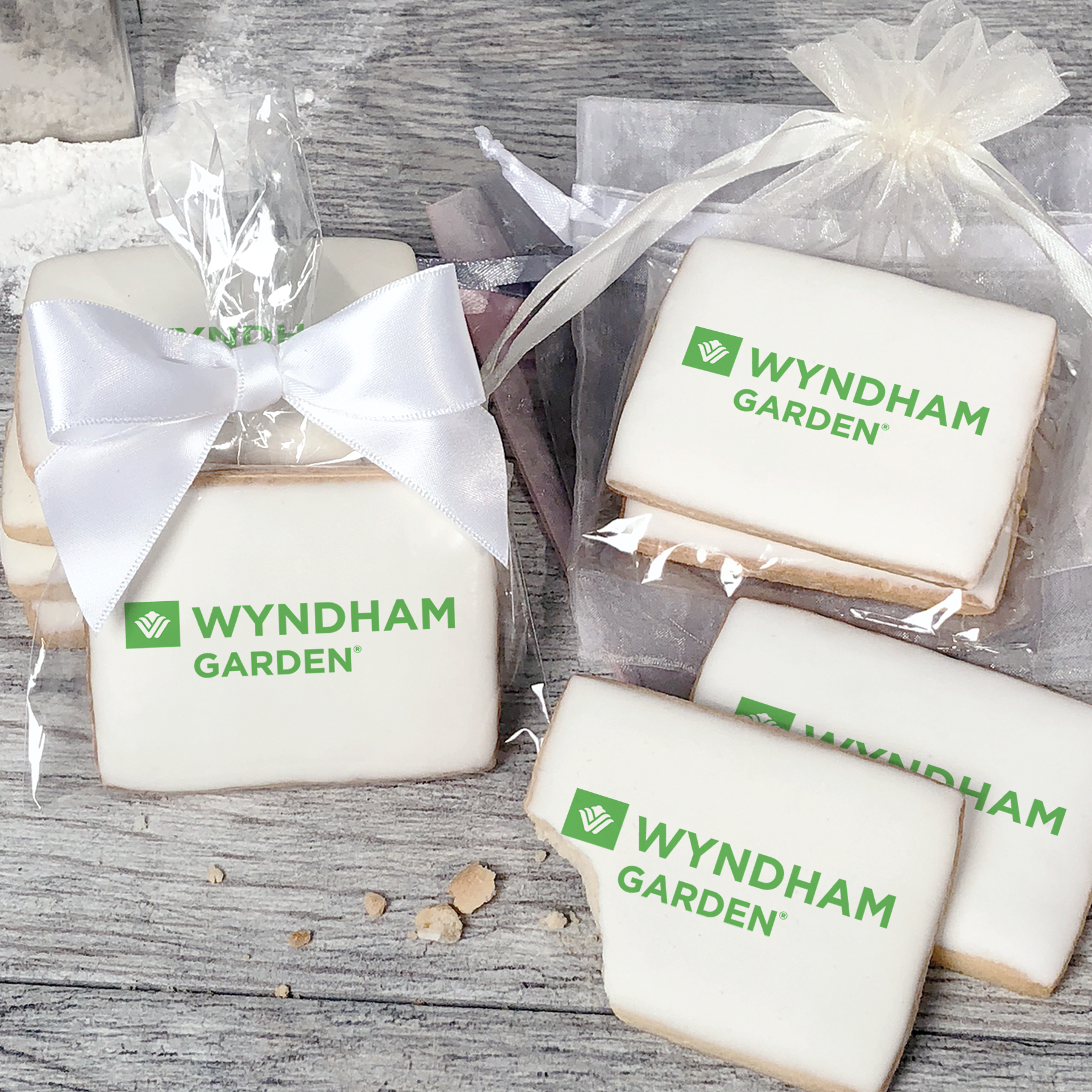 A stack of rectangular butter shortbread cookies with Wyndham Garden logo printed directly on a white, light sugar icing. Some cookies are shown in clear packaging with a twist-tie ribbon bow or inside an organza bag.