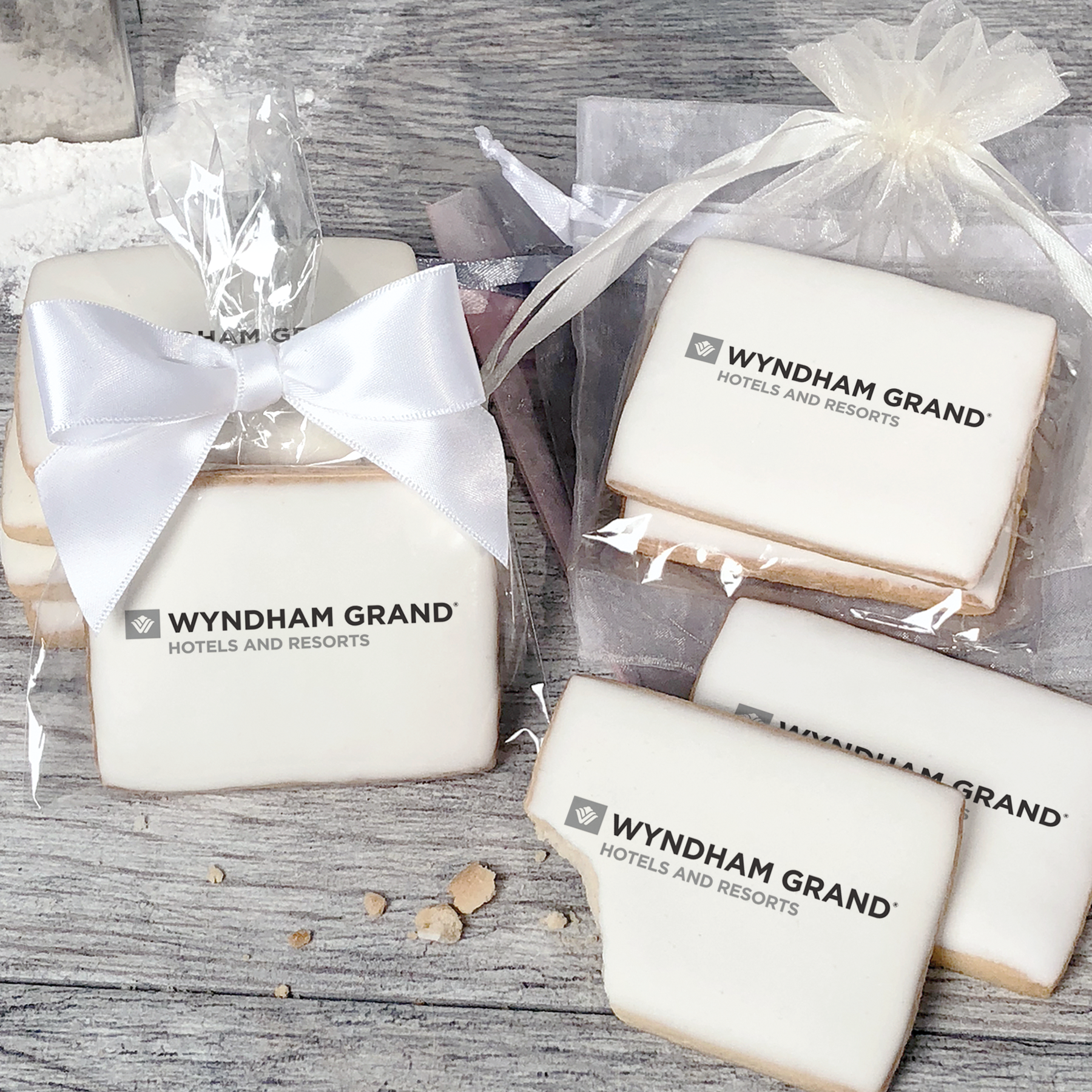 A stack of rectangular butter shortbread cookies with Wyndham Grand logo printed directly on a white, light sugar icing. Some cookies are shown in clear packaging with a twist-tie ribbon bow or inside an organza bag.