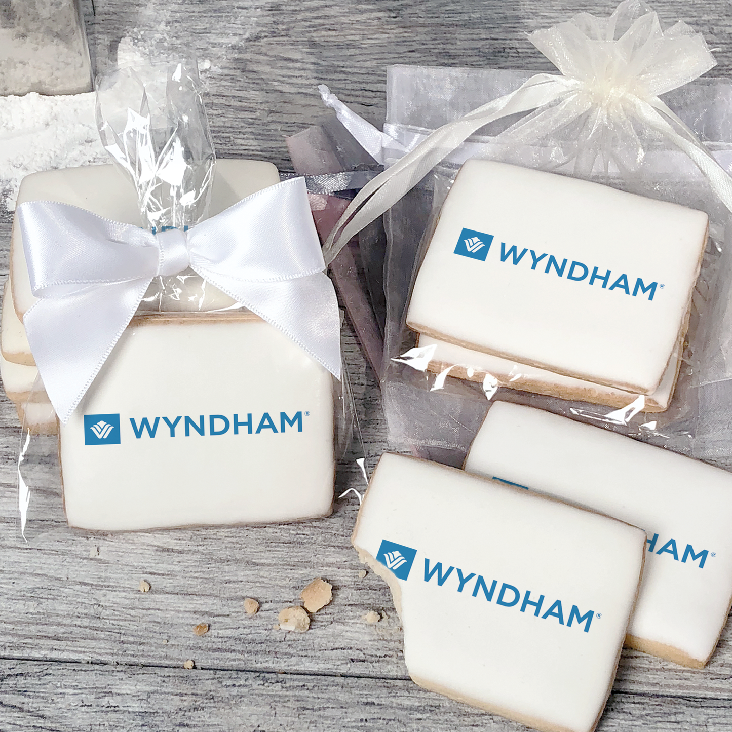 A stack of rectangular butter shortbread cookies with Wyndham logo printed directly on a white, light sugar icing. Some cookies are shown in clear packaging with a twist-tie ribbon bow or inside an organza bag.