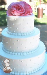 3-tier white cake with smooth white icing and decorated with white buttercream dots and white beaded trims and pink roses on top.