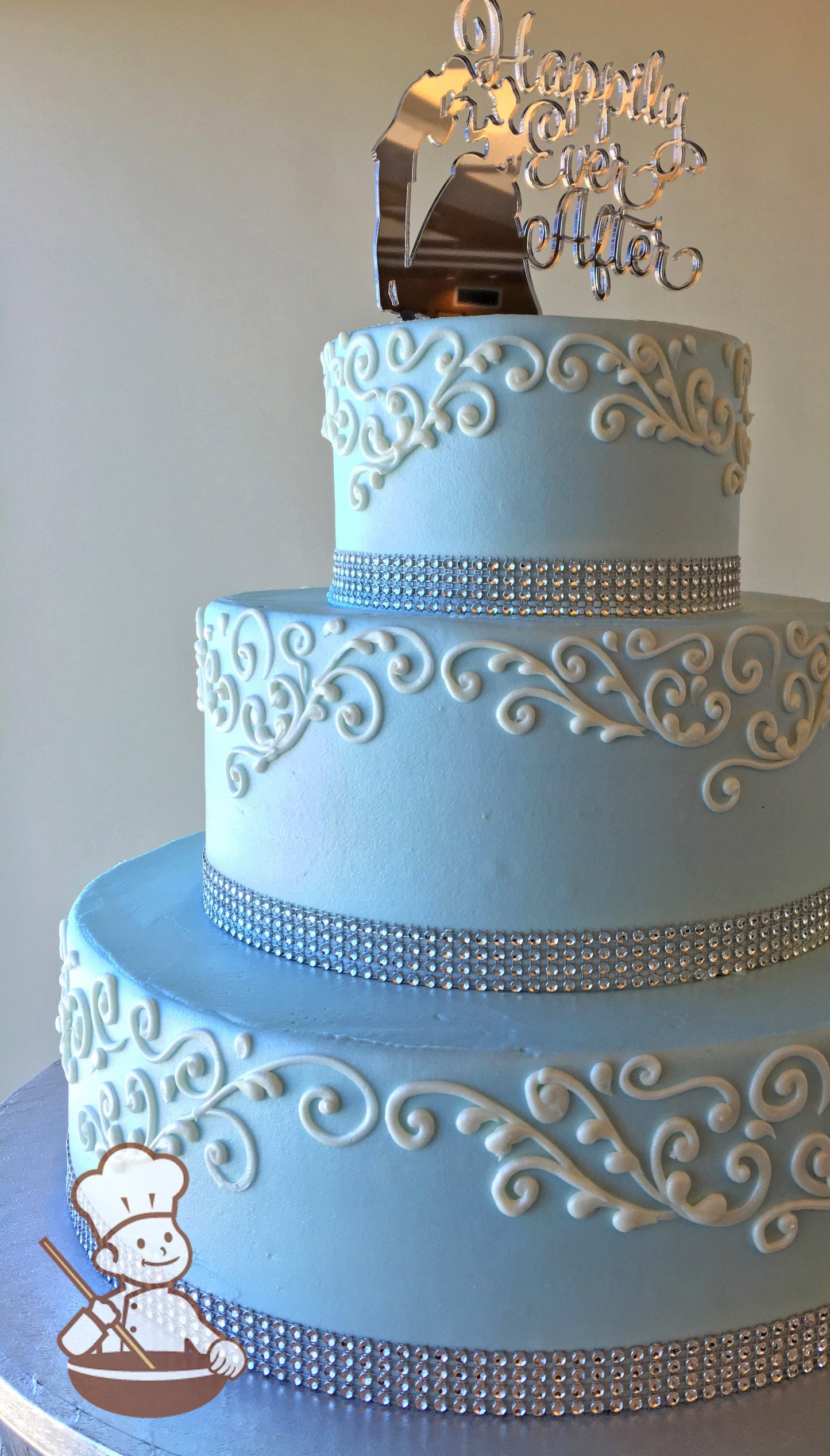 3-tier cake with smooth icing and decorated with white buttercream scrolls and a rhinestone band on the base of each tier.